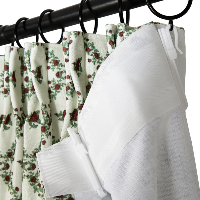 Close-up of pleated white curtains featuring a vibrant red rose and green leaf pattern, alongside a textured white liner, all suspended from a black curtain rod by black rings and white plastic hooks