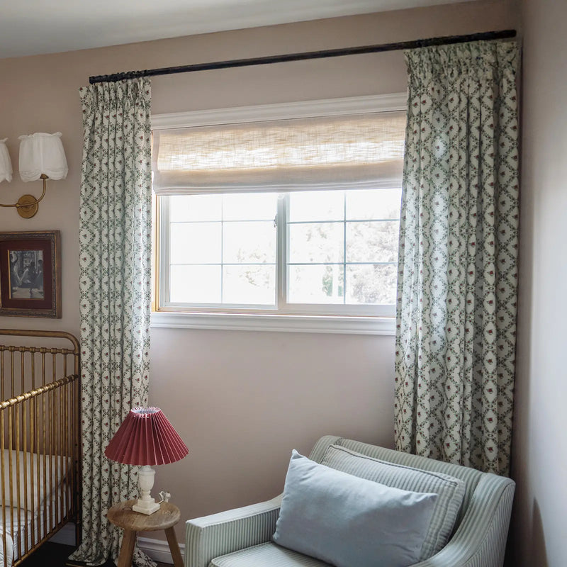 Interior of a charming nursery with a gold brass baby crib, a window dressed with sheer woven Roman shades and floral patterned curtains, and a light blue striped armchair next to a wooden stool topped with a pleated red lampshade