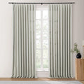Photograph of light grey linen blend curtains in a bright room, hanging from a black rod and framing a large window with white muntins and green trees visible outside, next to a fluted wooden accent table with a black vase of greenery and a brass candlestick