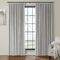 Light grey velvet pinch pleat curtains on a black rod frame a grid window with blurred foliage view, alongside an abstract tree painting and a beige armchair with a fringed throw blanket in a room