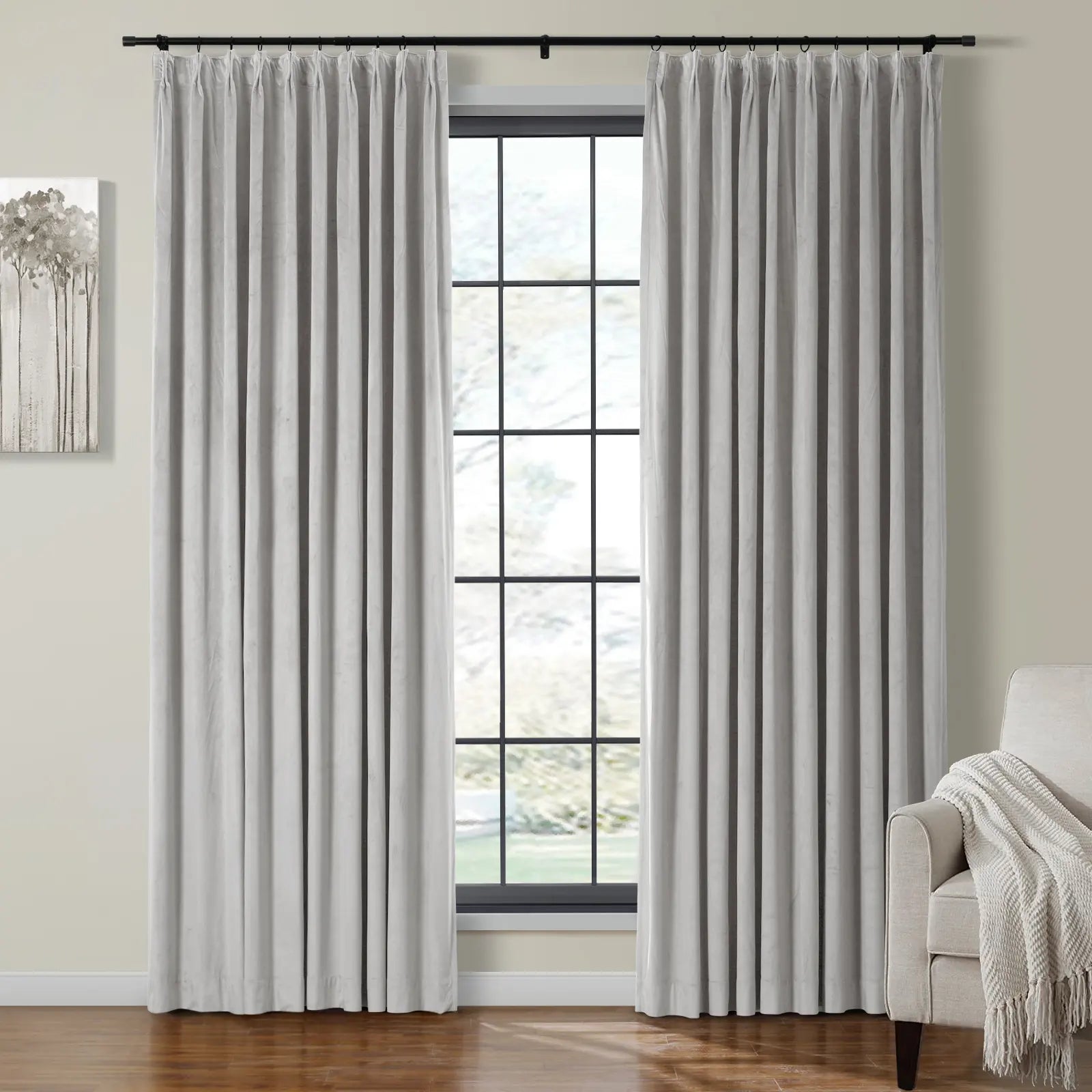 Light grey velvet pinch pleat curtains on a black rod frame a grid window with blurred foliage view, alongside an abstract tree painting and a beige armchair with a fringed throw blanket in a room