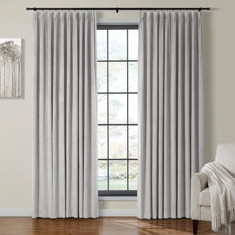 Light grey velvet pinch pleat curtains on a black rod frame a grid window with blurred foliage view, alongside an abstract tree painting and a beige armchair with a fringed throw blanket in a room