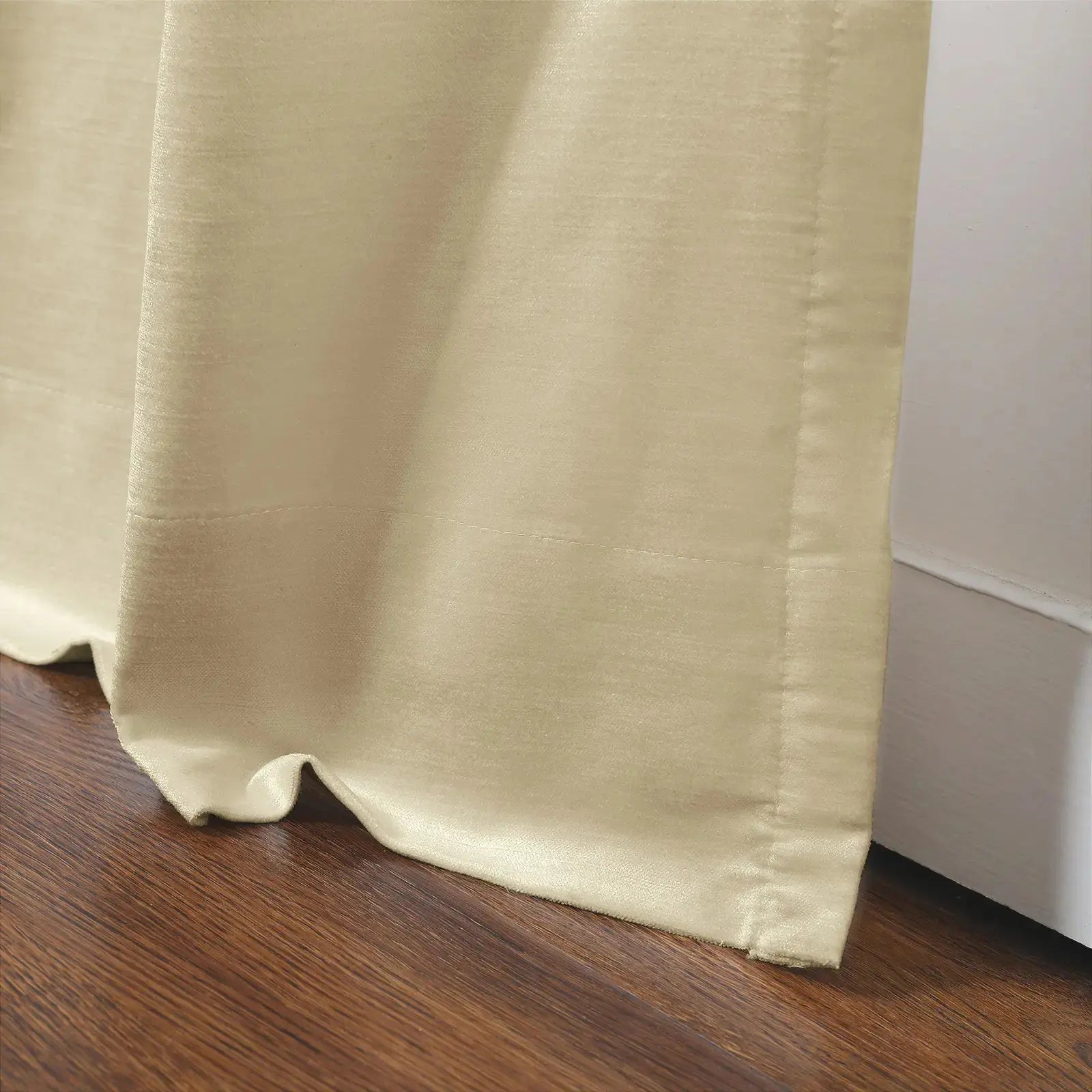 Cream curtain with a subtle sheen draped on a dark brown wooden floor next to a white baseboard with visible stitched details