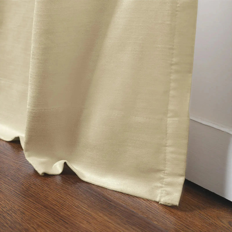Cream curtain with a subtle sheen draped on a dark brown wooden floor next to a white baseboard with visible stitched details