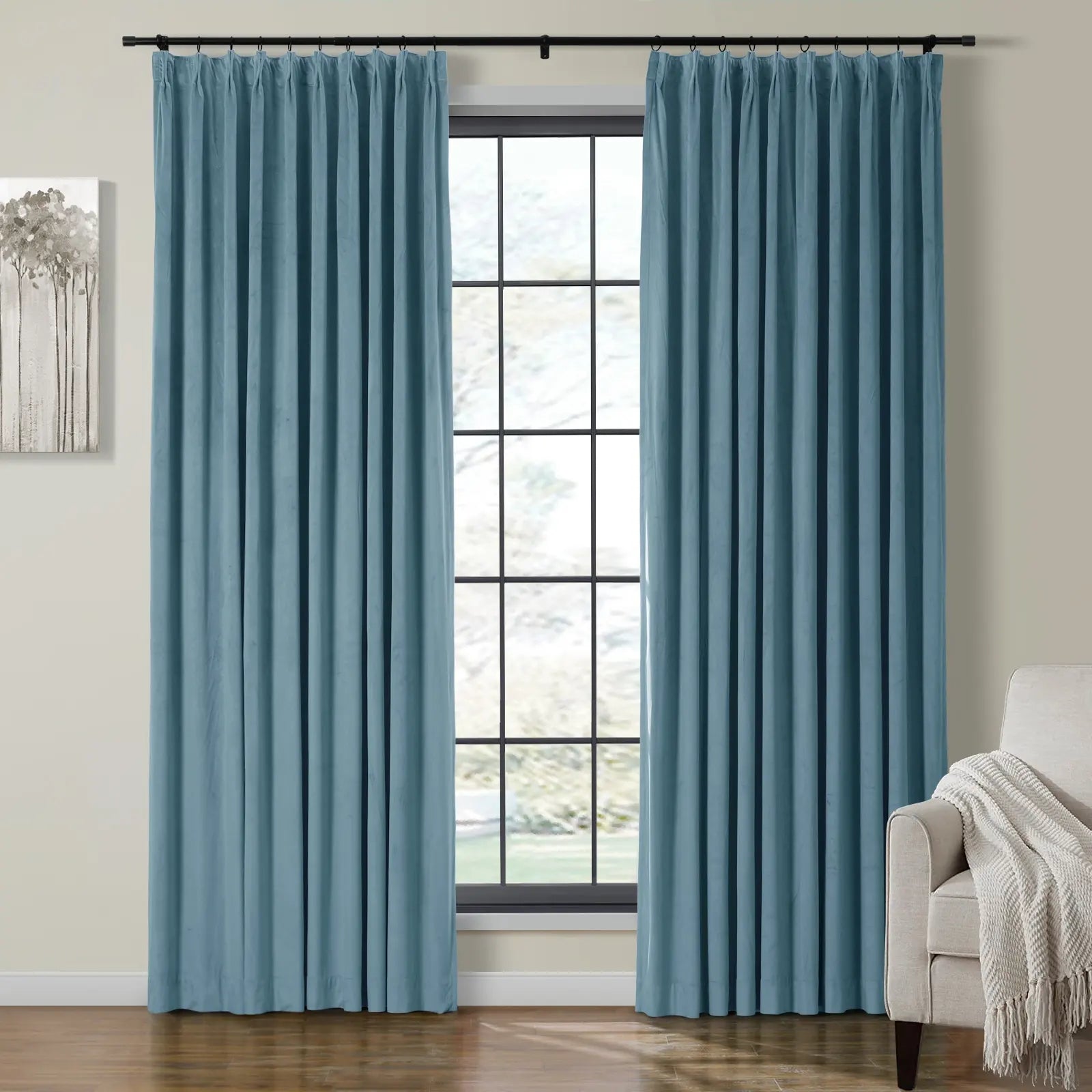 Soft blue pleated curtains on a black rod frame a grid-pane window overlooking a blurry outdoor scene, flanked by a minimalist tree painting and a cream armchair with a textured white throw