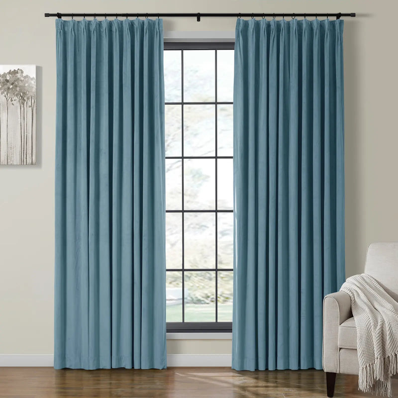 Soft blue pleated curtains on a black rod frame a grid-pane window overlooking a blurry outdoor scene, flanked by a minimalist tree painting and a cream armchair with a textured white throw