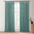 Teal textured linen pleated curtains on a black rod in a bright room framing a window revealing outdoor greenery beside a fluted wooden side table holding a black vase with a plant and a brass candle holder