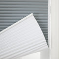 A window featuring layered window treatments with a gray cellular Blind in the background and a white pleated cellular Blind partially lowered diagonally into bright natural light