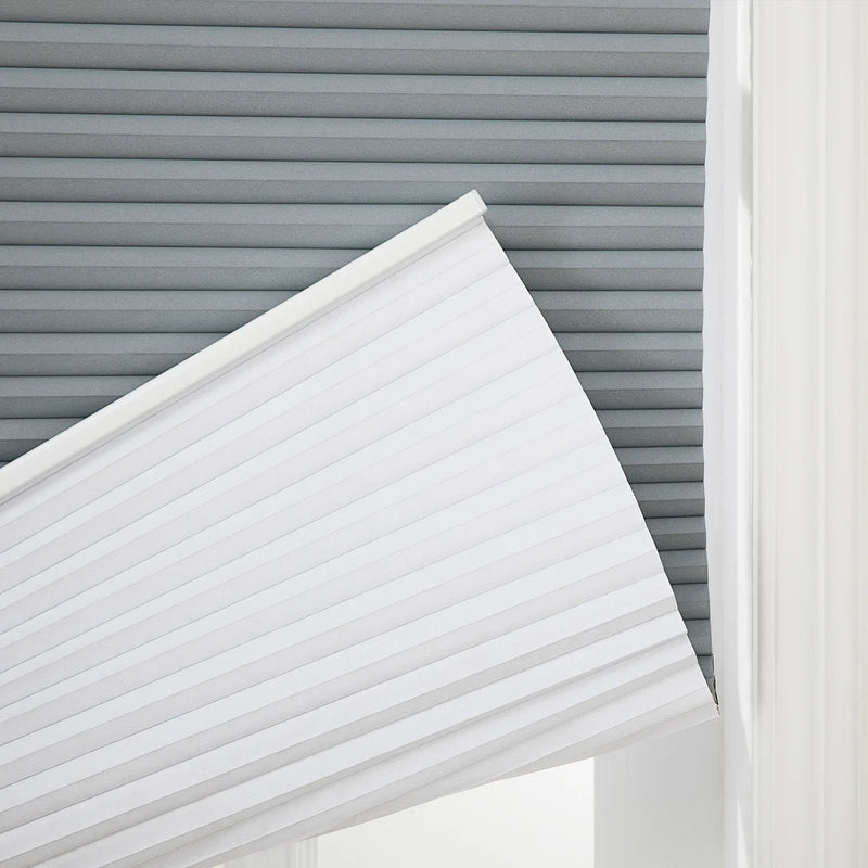 A window featuring layered window treatments with a gray cellular Blind in the background and a white pleated cellular Blind partially lowered diagonally into bright natural light