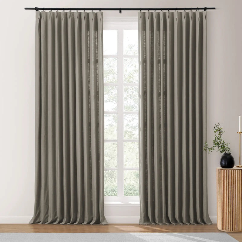 A modern interior features a pair of medium grey linen draperies on a black rod next to a white-framed window looking out to green trees, with a fluted wood side table holding a black vase of greenery and a gold candle holder
