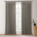 A modern interior features a pair of medium grey linen draperies on a black rod next to a white-framed window looking out to green trees, with a fluted wood side table holding a black vase of greenery and a gold candle holder