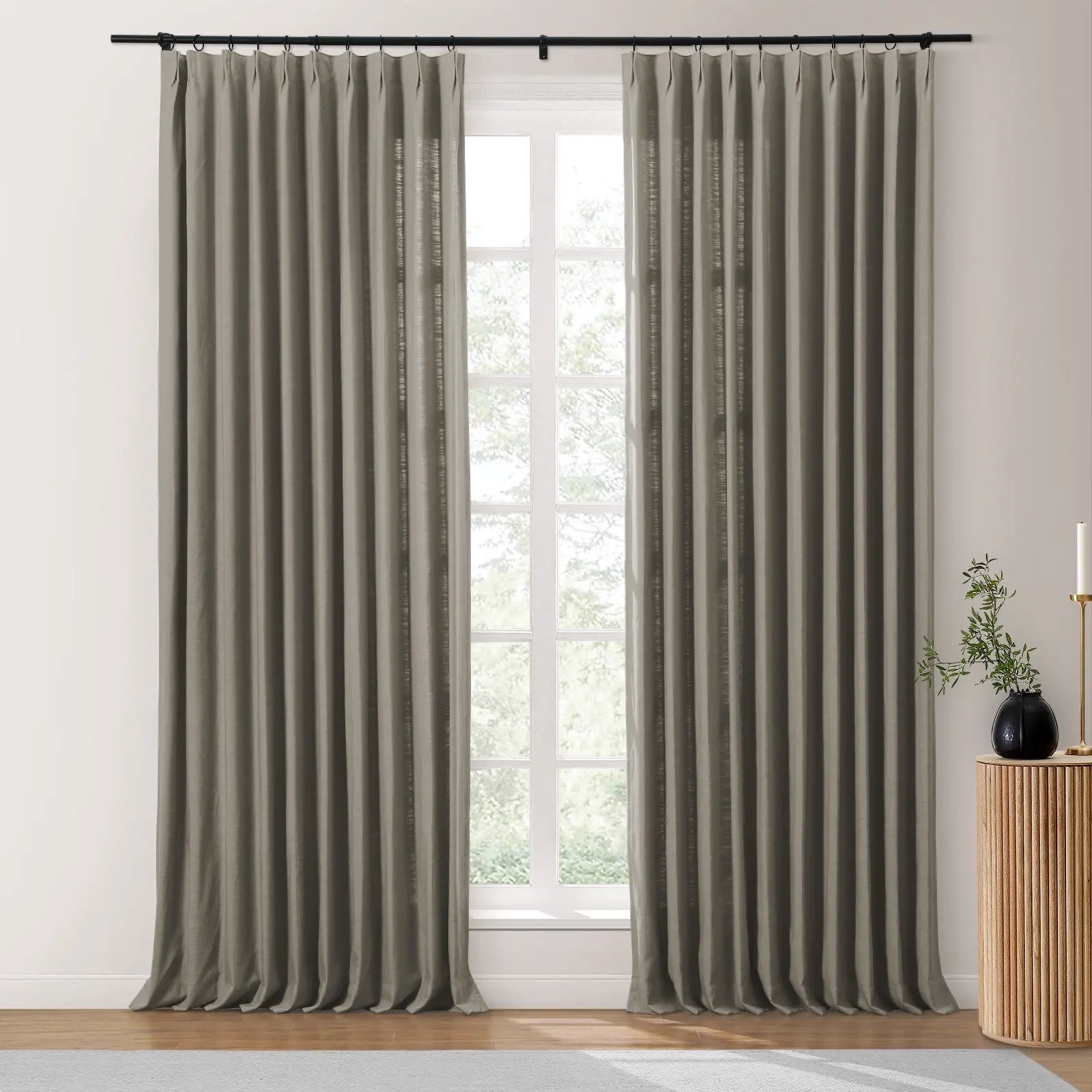 A modern interior features a pair of medium grey linen draperies on a black rod next to a white-framed window looking out to green trees, with a fluted wood side table holding a black vase of greenery and a gold candle holder