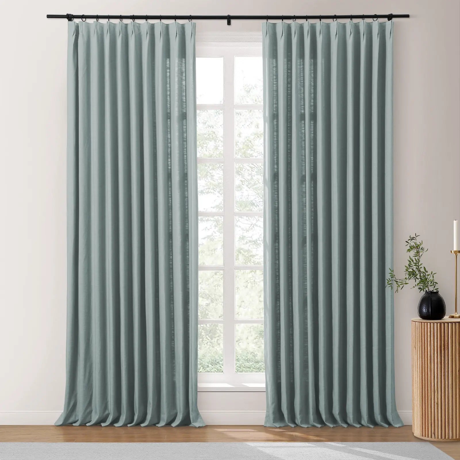 A pair of light sage green linen curtains on a black rod frame a window looking out to green trees, with a fluted wooden side table, black vase with green sprigs, and a gold candle holder to the right