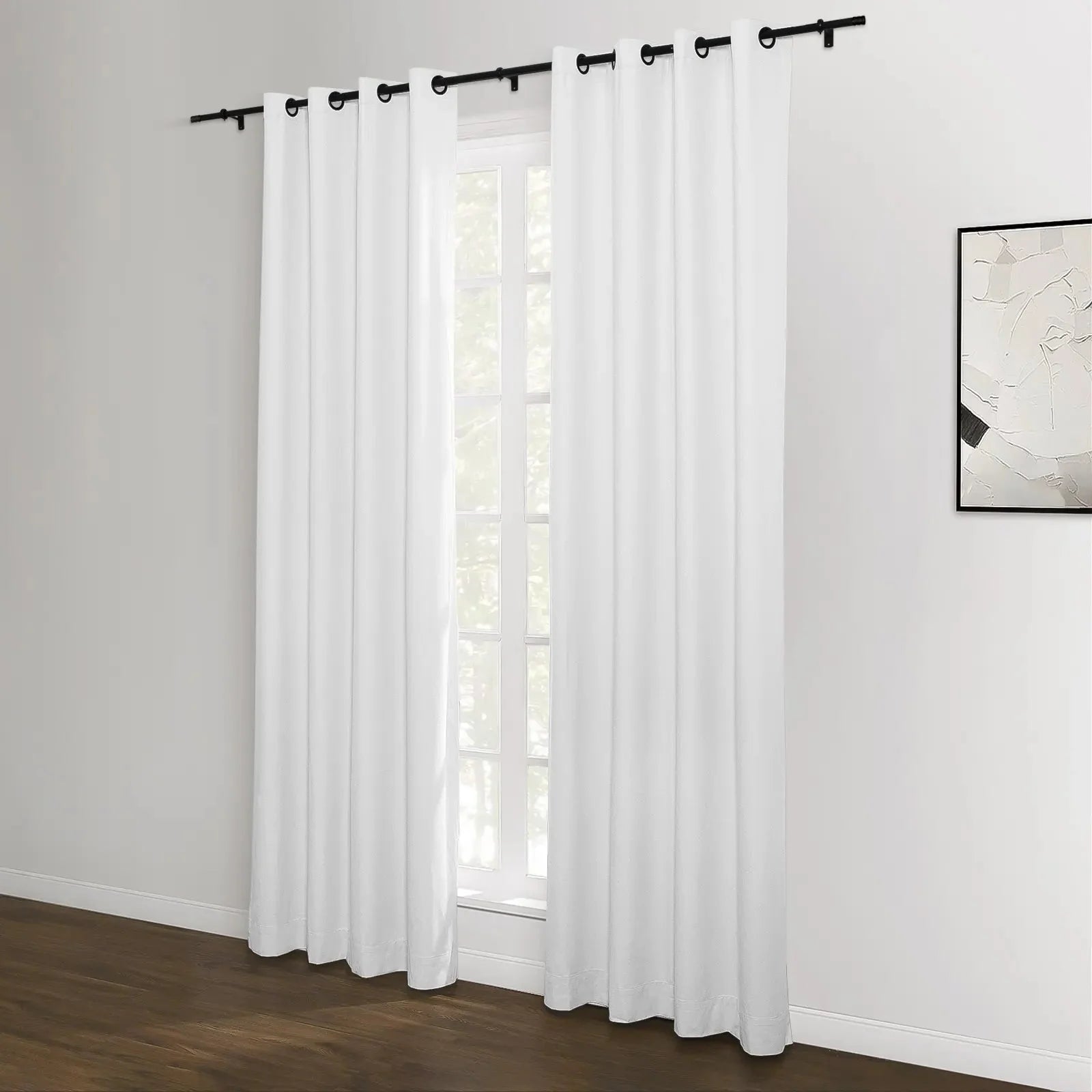 Photograph of a bright room featuring white grommet-top curtains on a black rod covering a window with a dark wood floor and a framed abstract black and white artwork on the right wall