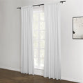 White pleated curtains on black rods frame a multi-pane window revealing green trees outside, set in a minimalist room with white walls, dark wood flooring, and a monochrome abstract artwork