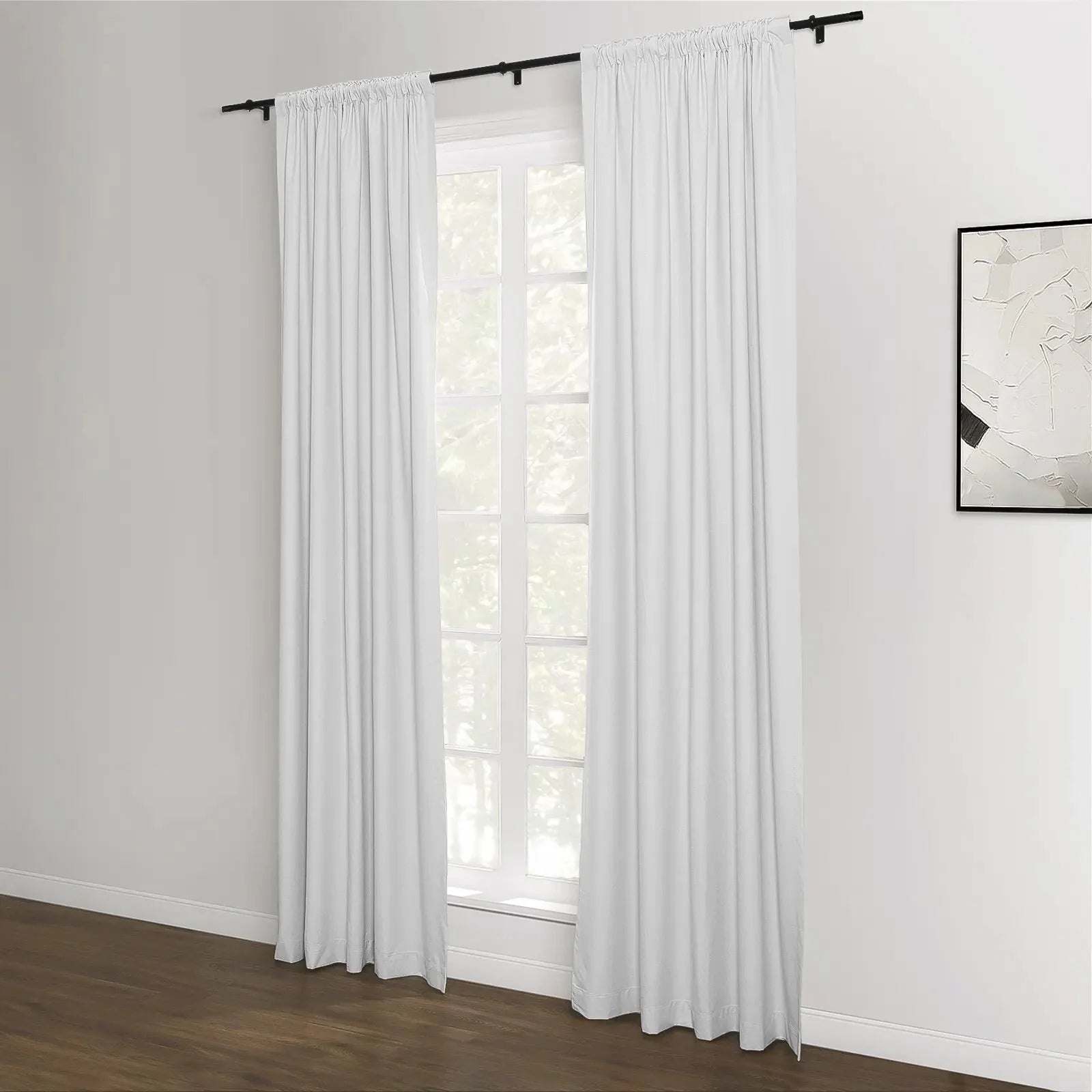 White pleated curtains on black rods frame a multi-pane window revealing green trees outside, set in a minimalist room with white walls, dark wood flooring, and a monochrome abstract artwork