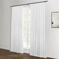 White pinch-pleat curtains on a black rod frame a large window with green foliage visible outside, complementing a modern room with dark wood flooring and abstract monochrome artwork