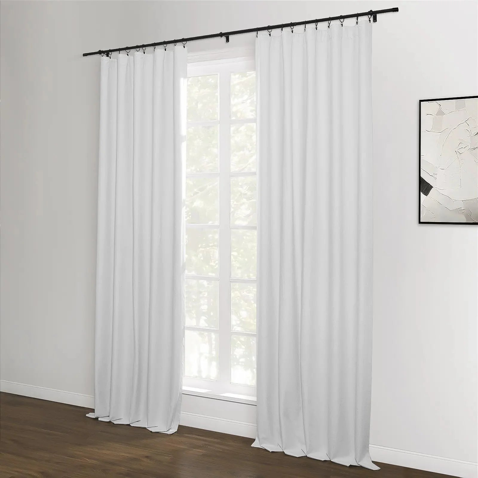 A room featuring white blackout curtains with a black rod and rings over a window, complemented by dark hardwood floors and an abstract black and white artwork