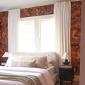 Photograph of a serene bedroom interior with a cream bouclé headboard, rich brown tree-patterned wallpaper, and bright white pleated curtains on a gold rod, flanked by dark fluted bedside tables