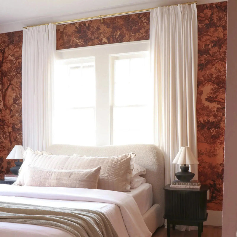 Photograph of a serene bedroom interior with a cream bouclé headboard, rich brown tree-patterned wallpaper, and bright white pleated curtains on a gold rod, flanked by dark fluted bedside tables