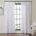 White pleated curtains hang on a black rod framing a large grid-paned window in a contemporary room featuring abstract tree artwork and a beige armchair with a cream throw
