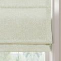 Photograph of a light green linen Roman blind featuring a subtle floral damask pattern and neat horizontal folds, framed by a white window jamb with a blurred green outdoor scene below