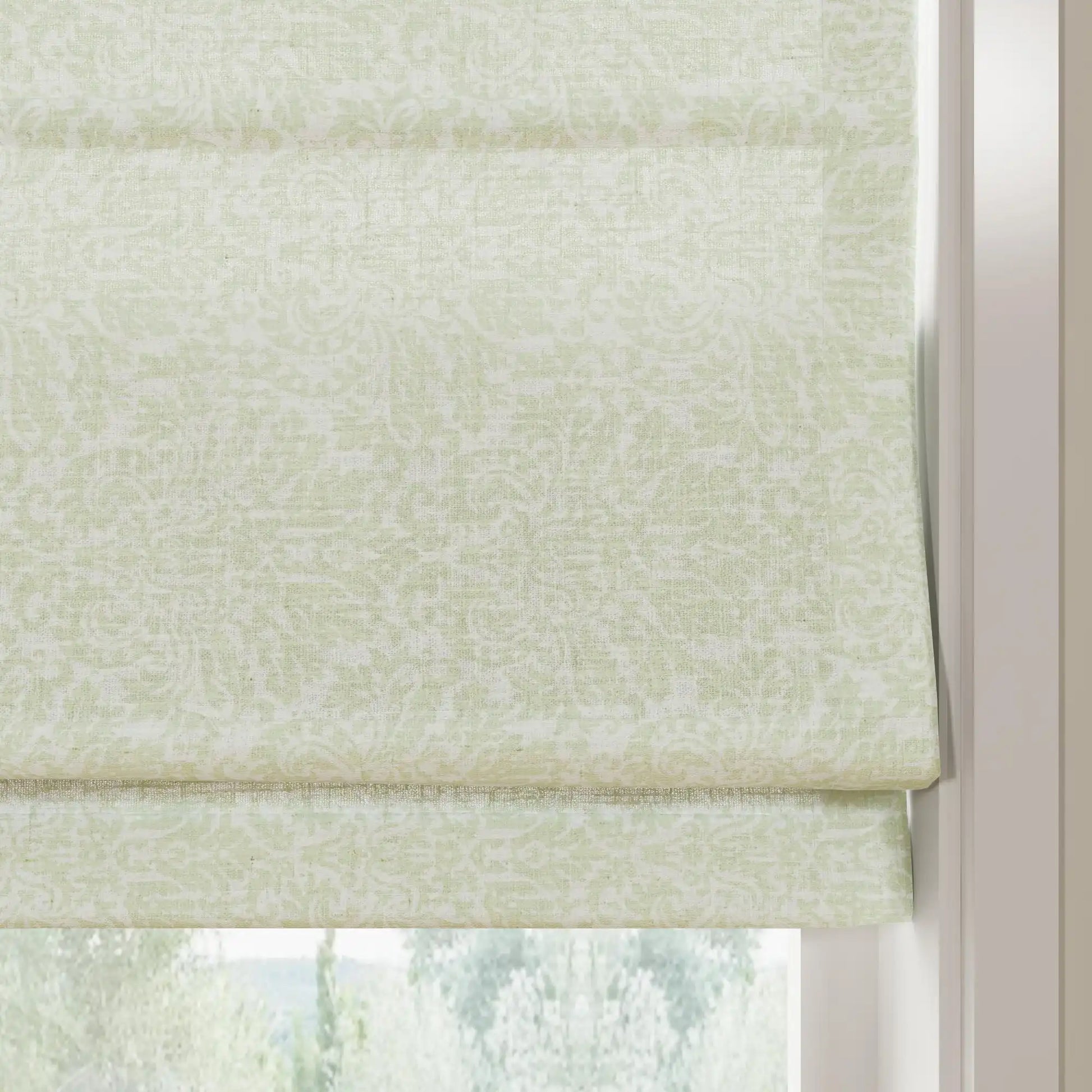 Photograph of a light green linen Roman blind featuring a subtle floral damask pattern and neat horizontal folds, framed by a white window jamb with a blurred green outdoor scene below
