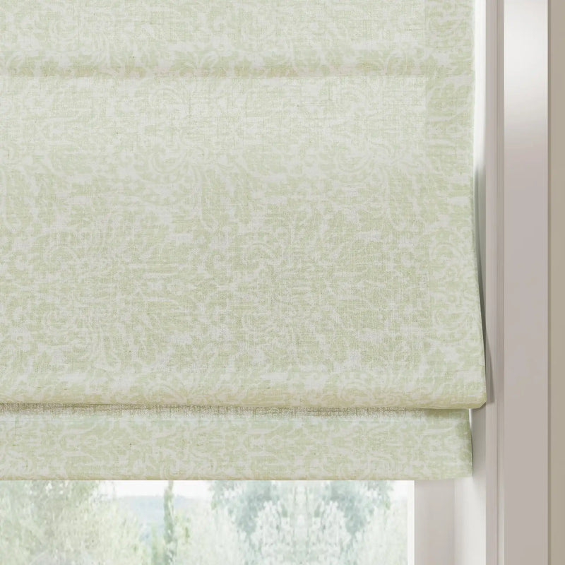 Photograph of a light green linen Roman blind featuring a subtle floral damask pattern and neat horizontal folds, framed by a white window jamb with a blurred green outdoor scene below