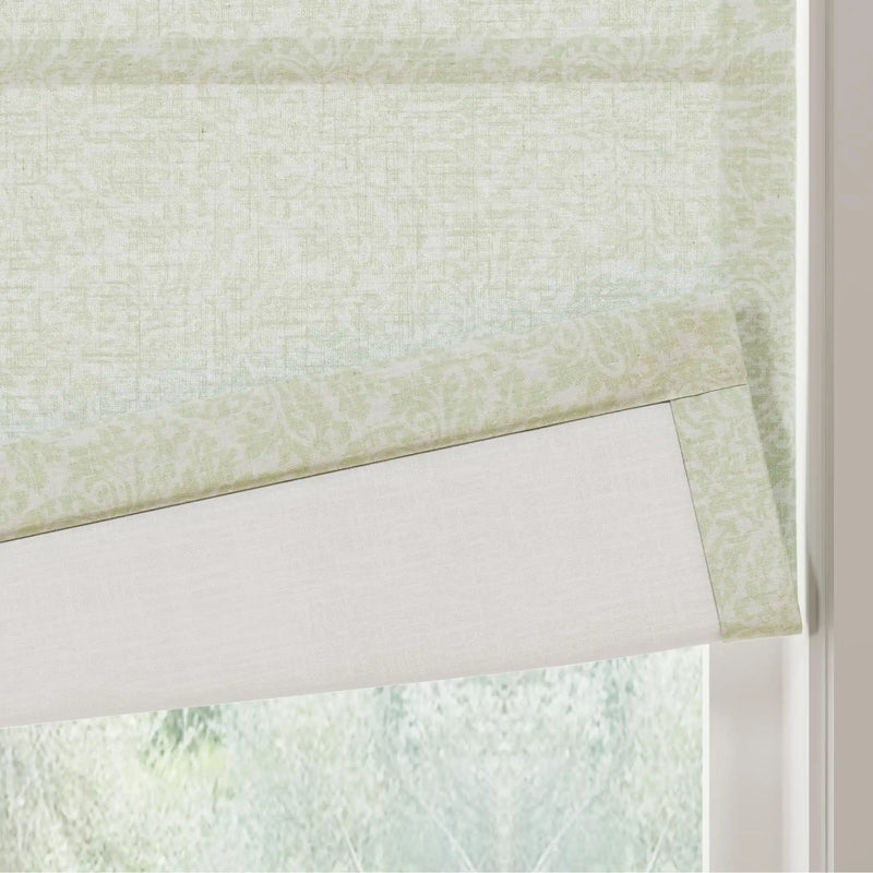 A light green patterned Roman shade featuring a subtle botanical damask print with a solid white lining partially lowered against a window
