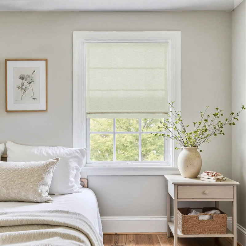 A neutral-toned bedroom interior featuring a window with a white Roman shade, a framed botanical print, and a light wood nightstand displaying a ceramic vase with green branches and a woven storage basket