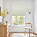 Bright interior room with a window featuring a cream roman shade overlooking green trees, a natural wood dresser with a white vase of vibrant yellow flowers and stacked books, and a matching wooden chair with a white cushion on a light rug
