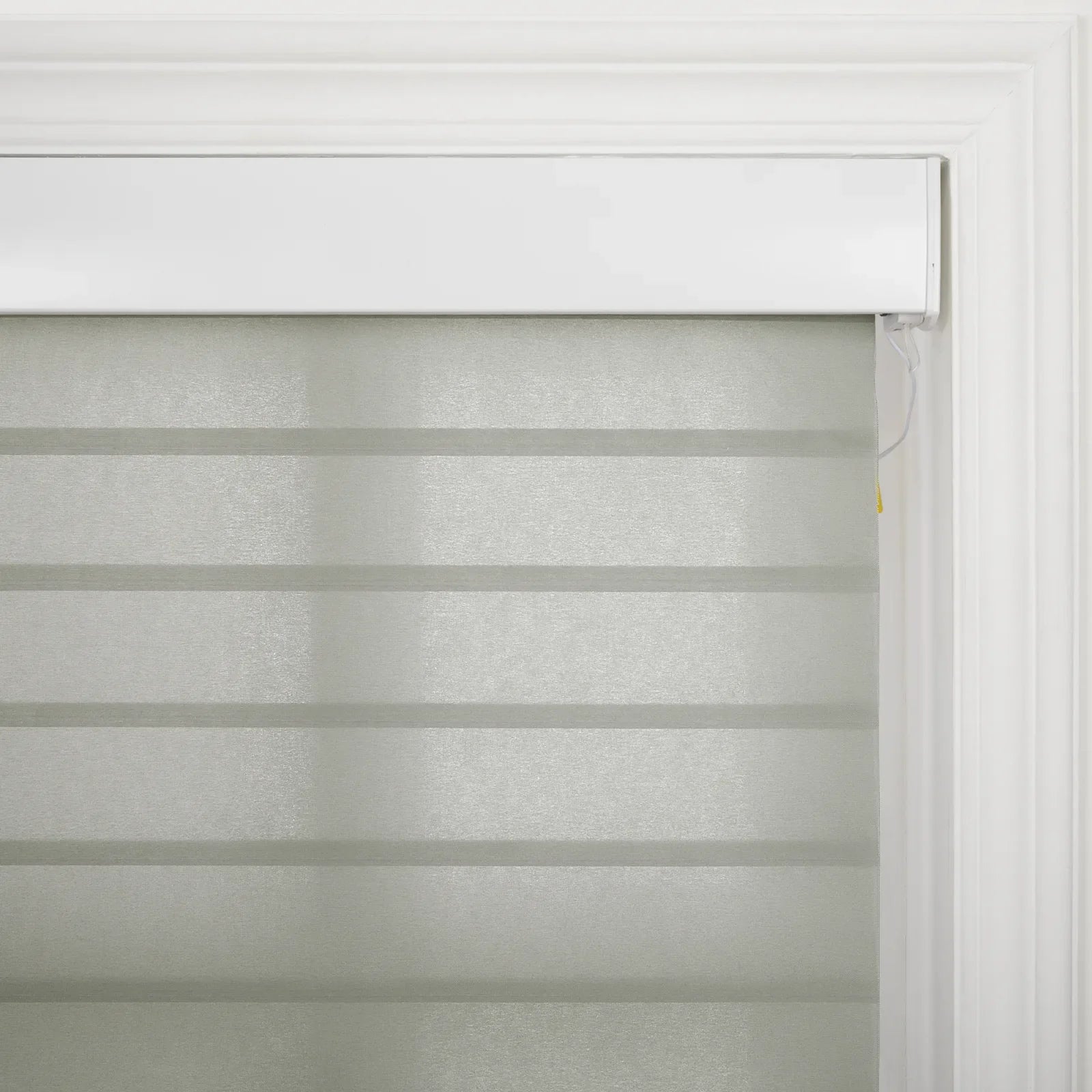 A light grey zebra sheer window Blind with alternating opaque and sheer horizontal stripes installed within white decorative window trim showing a subtle yellow tip on the control cord