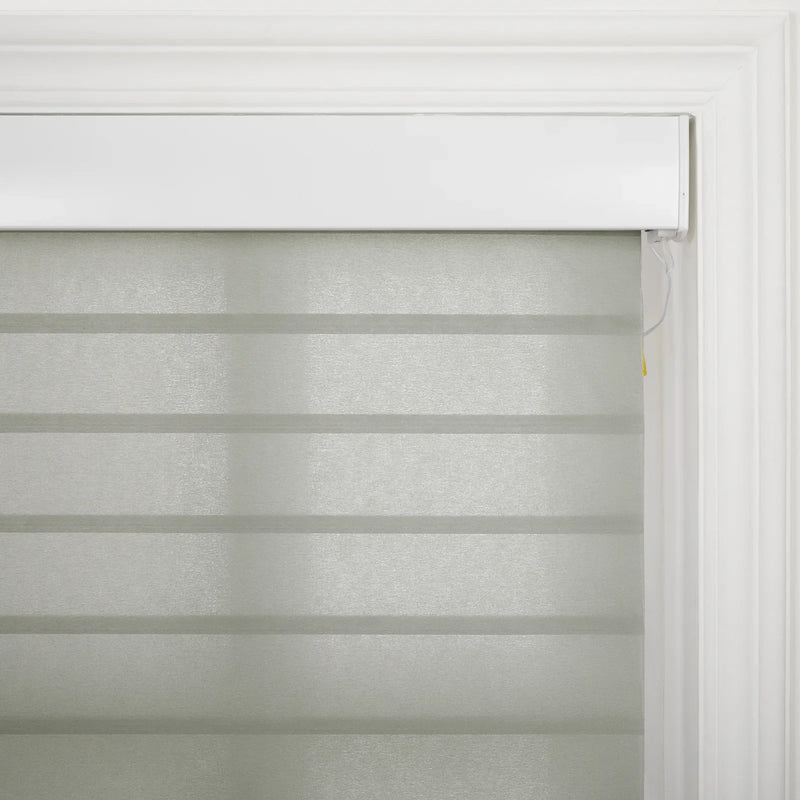 A light grey zebra sheer window Blind with alternating opaque and sheer horizontal stripes installed within white decorative window trim showing a subtle yellow tip on the control cord