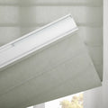 A light green pleated fabric window Blind partially lowered, showcasing its woven texture, white headrail, and a blurred view of outdoor greenery