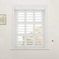 White layered zebra roller Blinds cover a window with white trim, flanked by a framed botanical art print on the left and a white remote control on the right, providing a filtered view of autumn trees