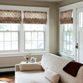 Bright living room interior featuring a light beige linen sofa with a brown throw, positioned before large windows adorned with patterned brown Roman shades, and a vase of dried flowers beside a framed photo on the armrest