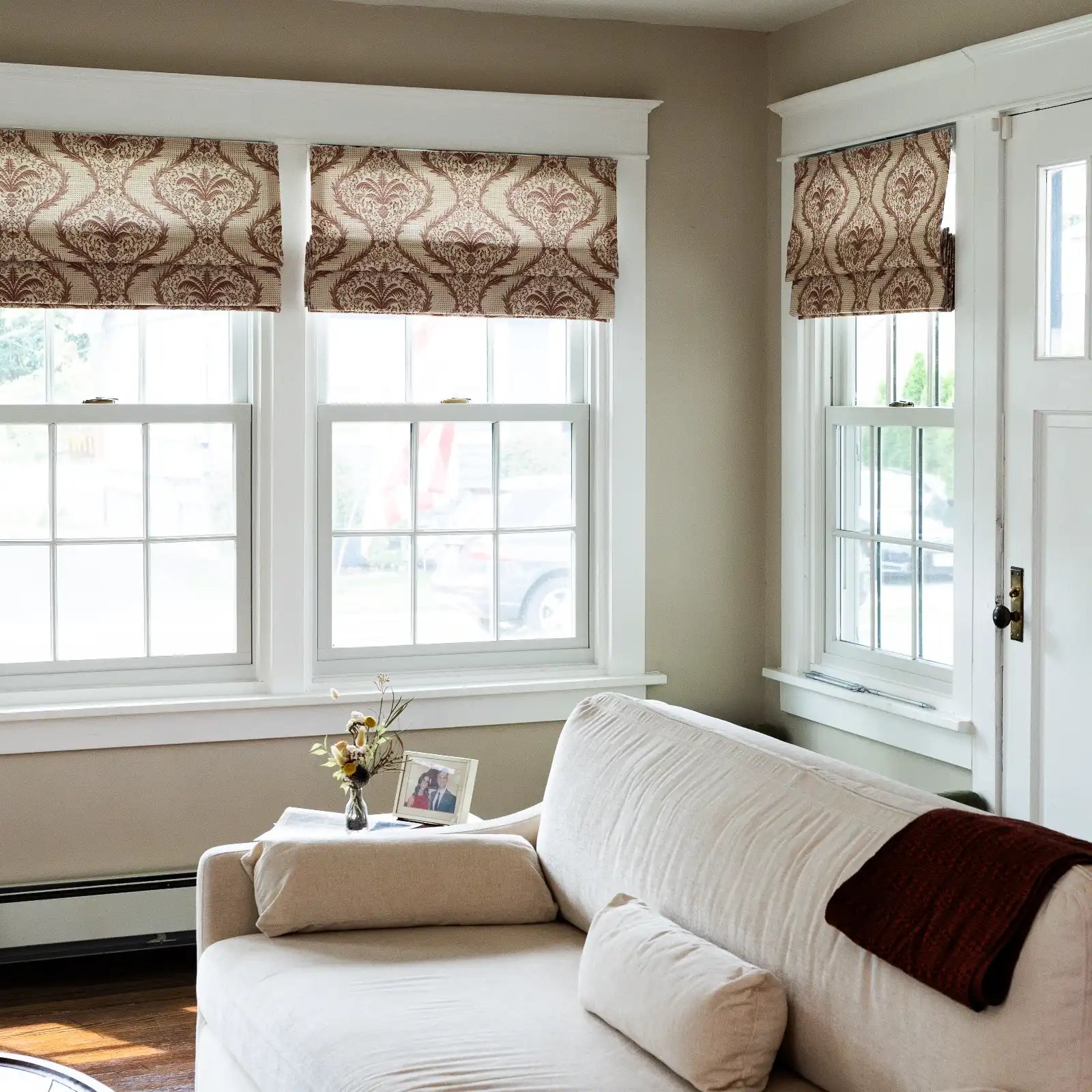 Bright living room interior featuring a light beige linen sofa with a brown throw, positioned before large windows adorned with patterned brown Roman shades, and a vase of dried flowers beside a framed photo on the armrest