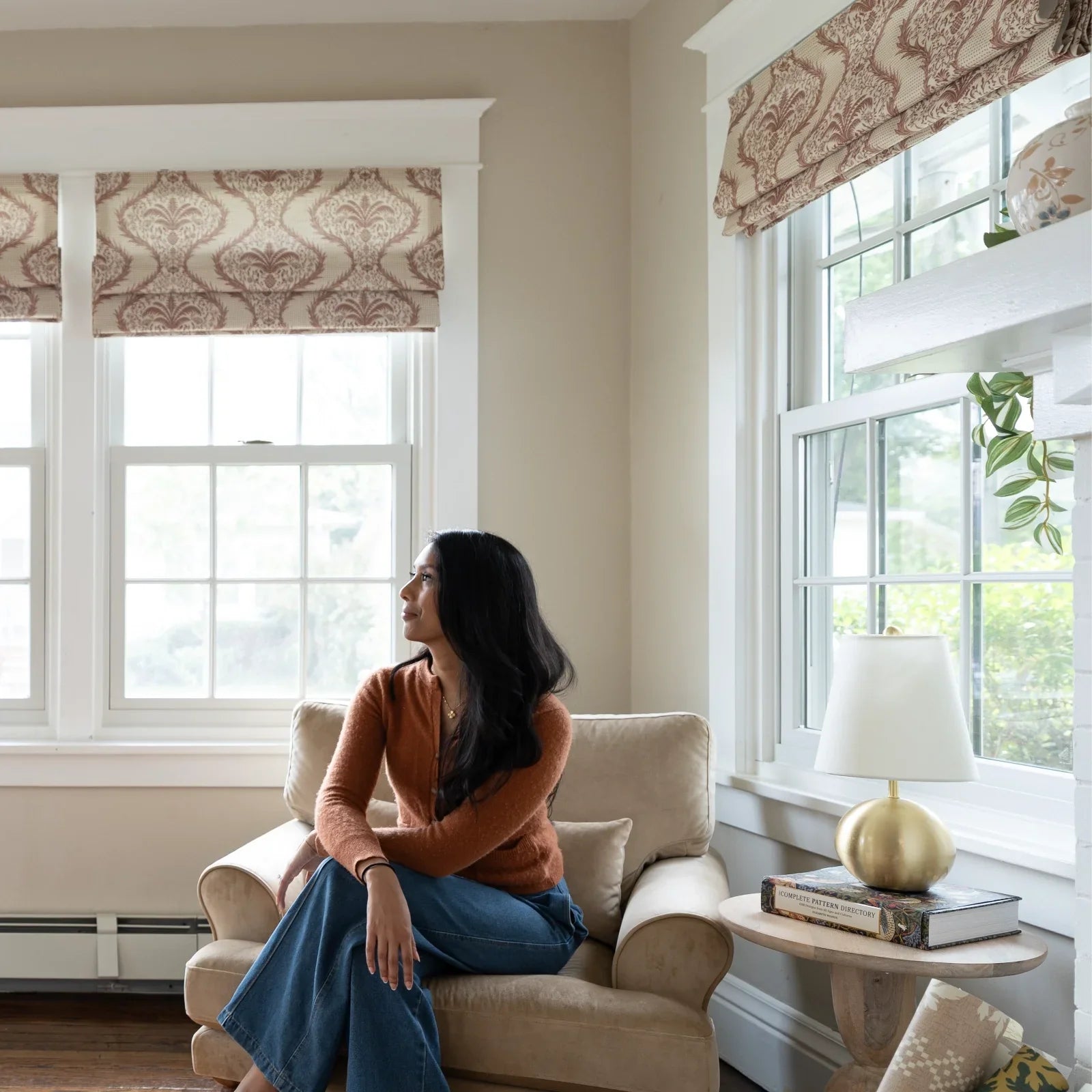 A woman with long dark hair sits in a beige armchair wearing an orange cardigan and blue wide-leg jeans looking out large windows framed by patterned roman blinds next to a side table with a gold spherical lamp and a trailing green plant