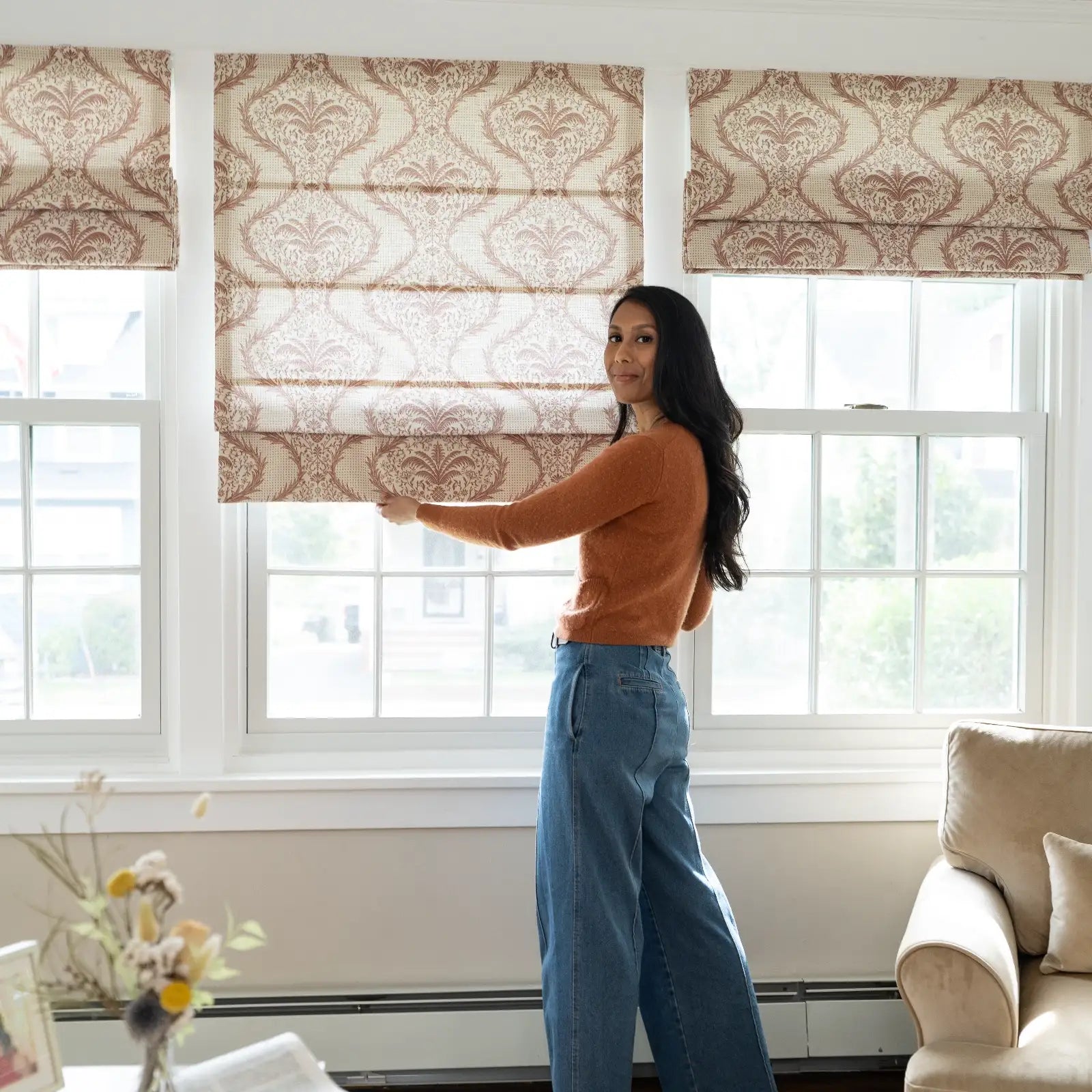 Photograph of a woman in an orange sweater and wide-leg jeans adjusting a brown damask patterned Roman shade on one of three white-framed windows in a bright living room with a beige armchair