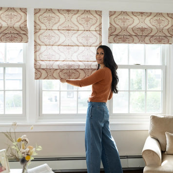 Photograph of a woman in an orange sweater and wide-leg jeans adjusting a brown damask patterned Roman shade on one of three white-framed windows in a bright living room with a beige armchair