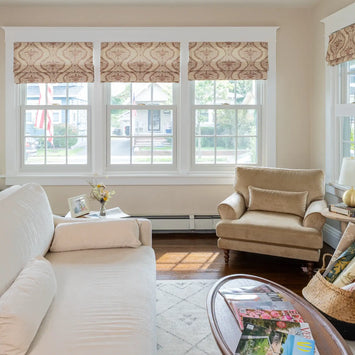 A bright residential living room interior featuring a cream sofa, beige armchair, patterned Roman shades, and magazines on an oval coffee table, with American flags visible through the windows