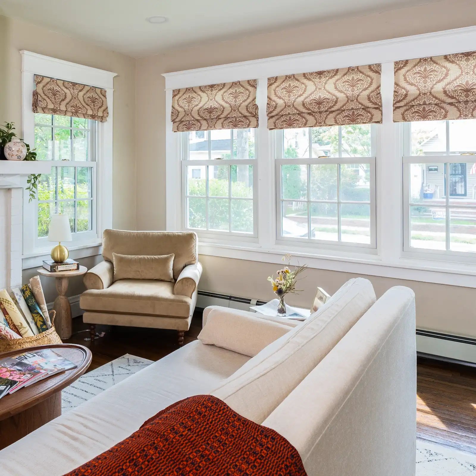 A cozy living room interior with a cream chaise lounge draped with an orange patterned throw, a beige armchair, large windows featuring brown patterned Roman blinds, and a distinctive gold spherical table lamp
