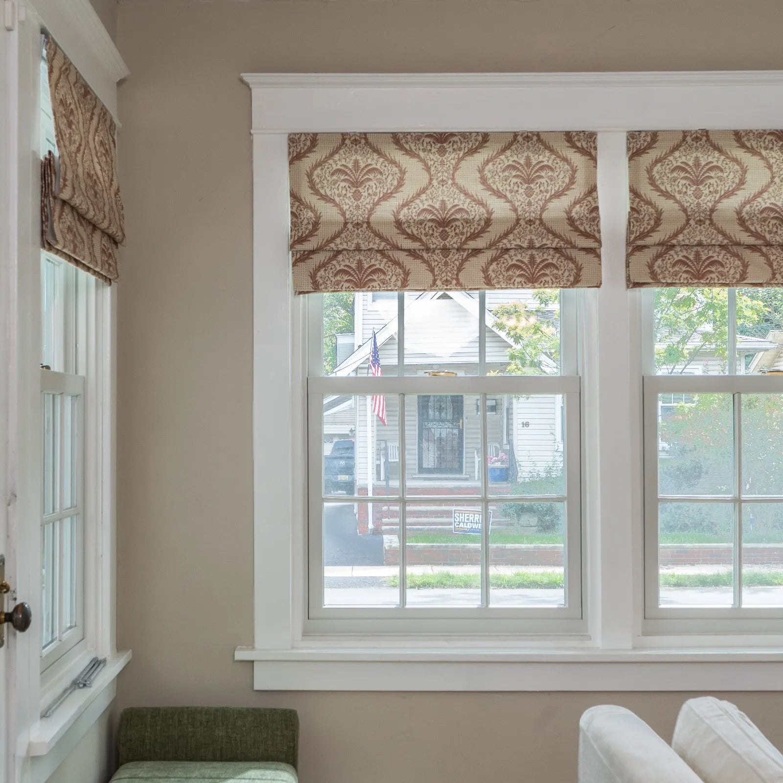 Photograph of an interior window with beige and brown damask patterned Roman shades looking out to a residential street with a white house, an American flag, and a Sherri Caldwell yard sign
