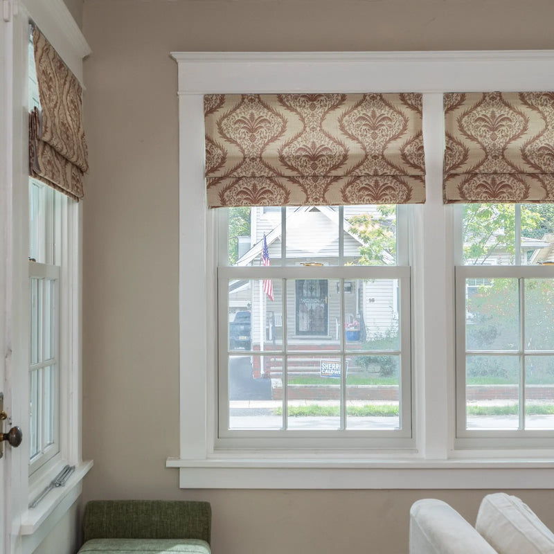 Photograph of an interior window with beige and brown damask patterned Roman shades looking out to a residential street with a white house, an American flag, and a Sherri Caldwell yard sign