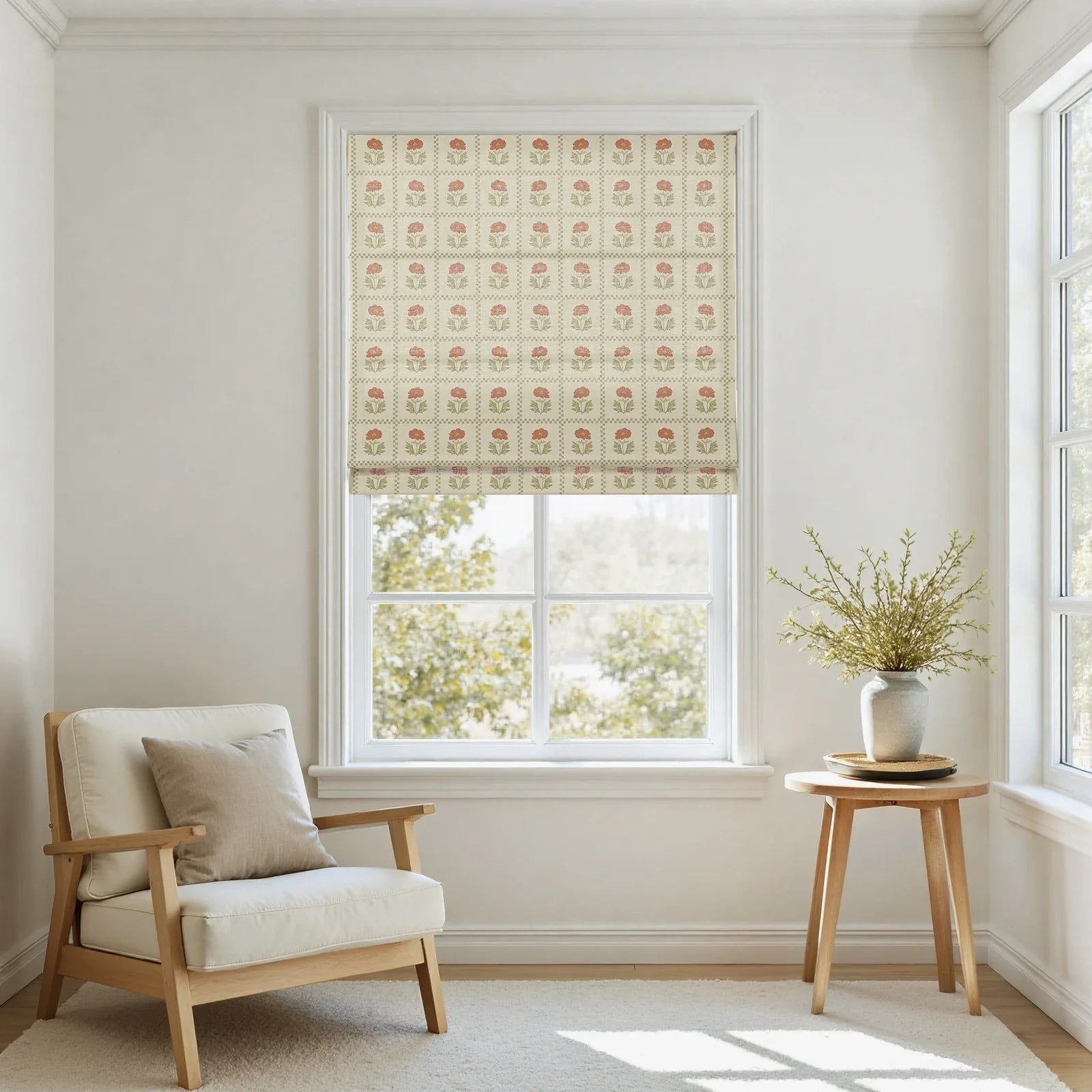 An airy interior room showcases a window with a cream Roman shade featuring a red floral block print pattern, a light wood armchair with cream cushions and a linen pillow, and a small round wooden side table holding a gray vase with green foliage