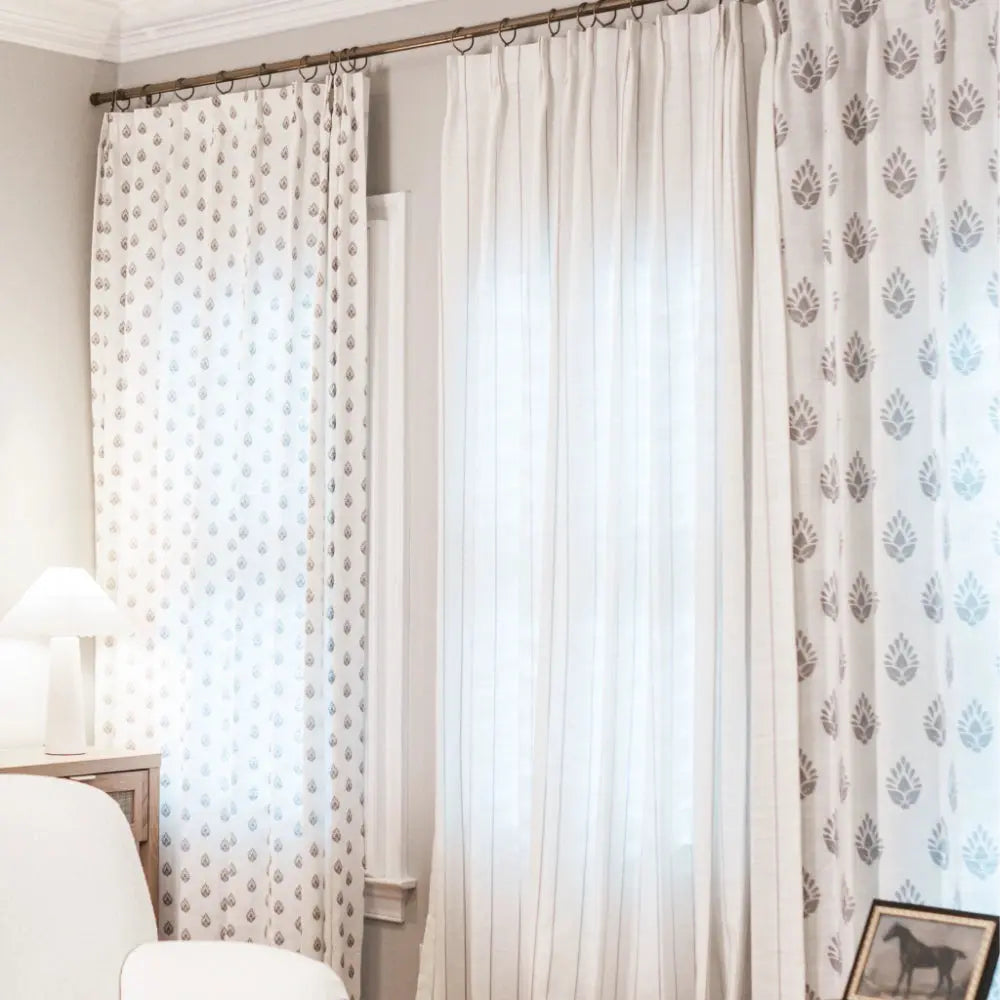 Photograph of an interior room featuring layered window treatments: white patterned curtains with gray teardrop and leaf designs, sheer striped white curtains, a bronze rod, a white table lamp, and a framed horse picture
