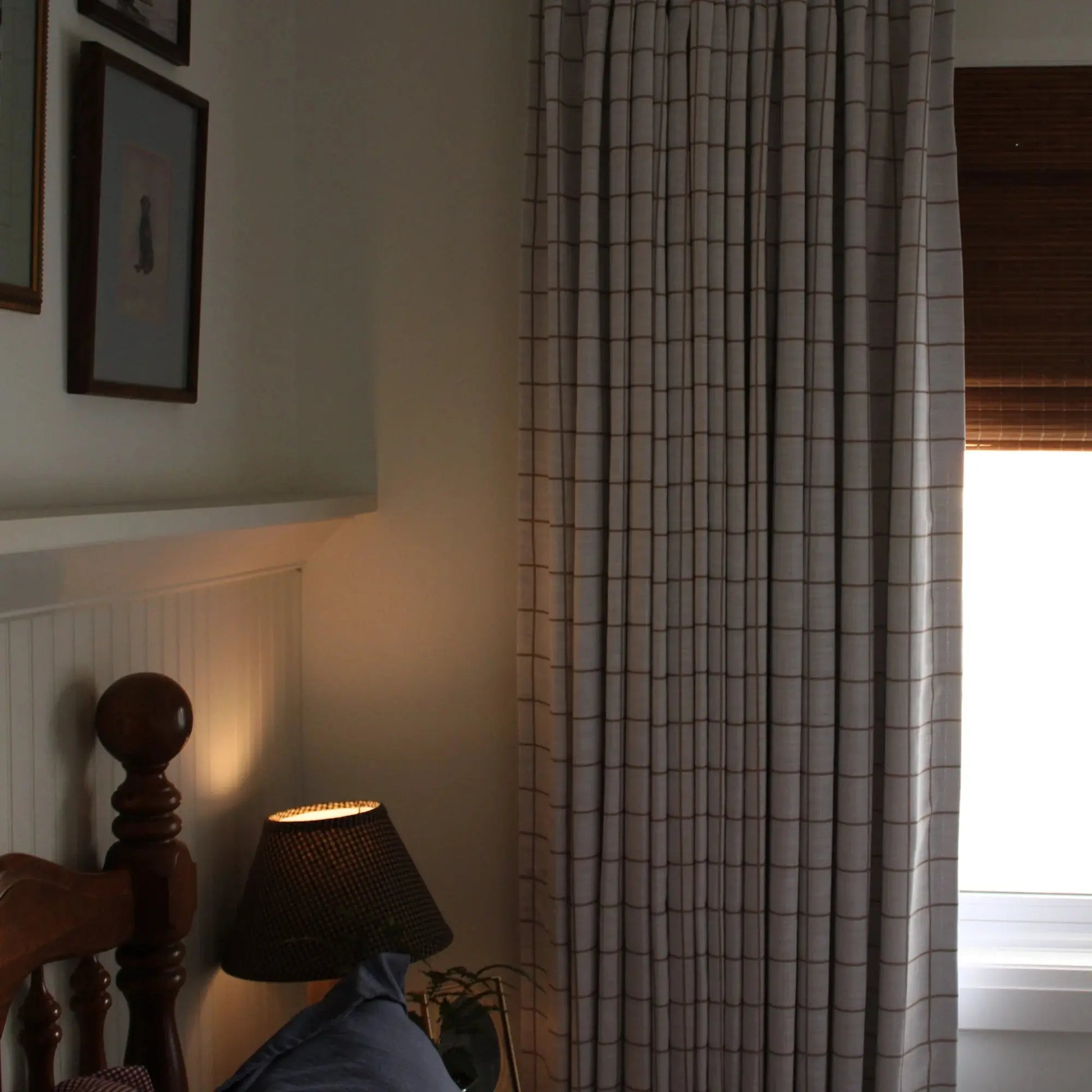 Photography of a cozy bedroom corner with a dark wooden bed frame and spherical finial, a warm glowing lamp with a woven shade, and light gray grid-patterned curtains over brown bamboo blinds