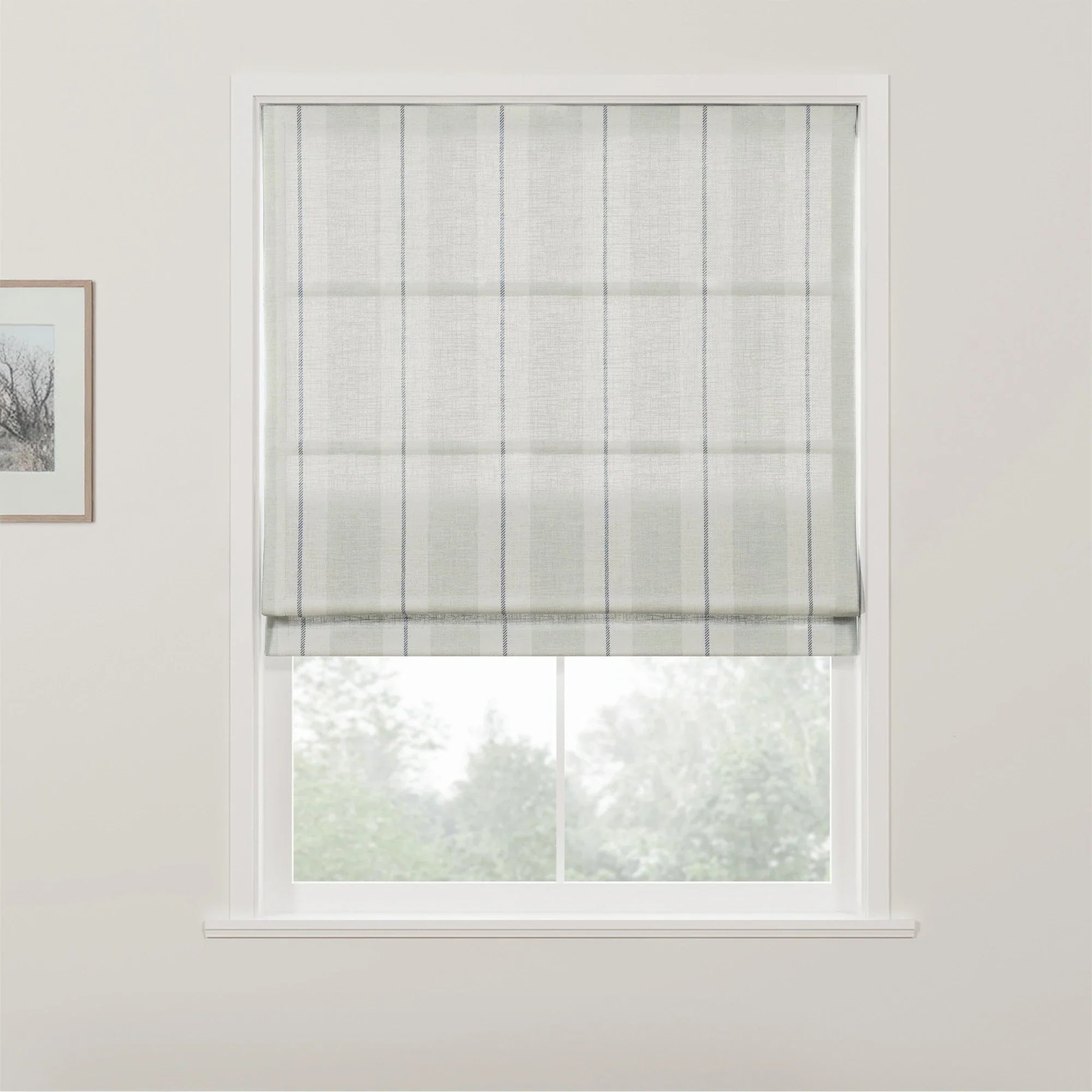 A light gray textured Roman blind with thin vertical blue stripes partially lowered in a white framed window offering a blurred view of green trees with a portion of a framed artwork visible on the left interior wall