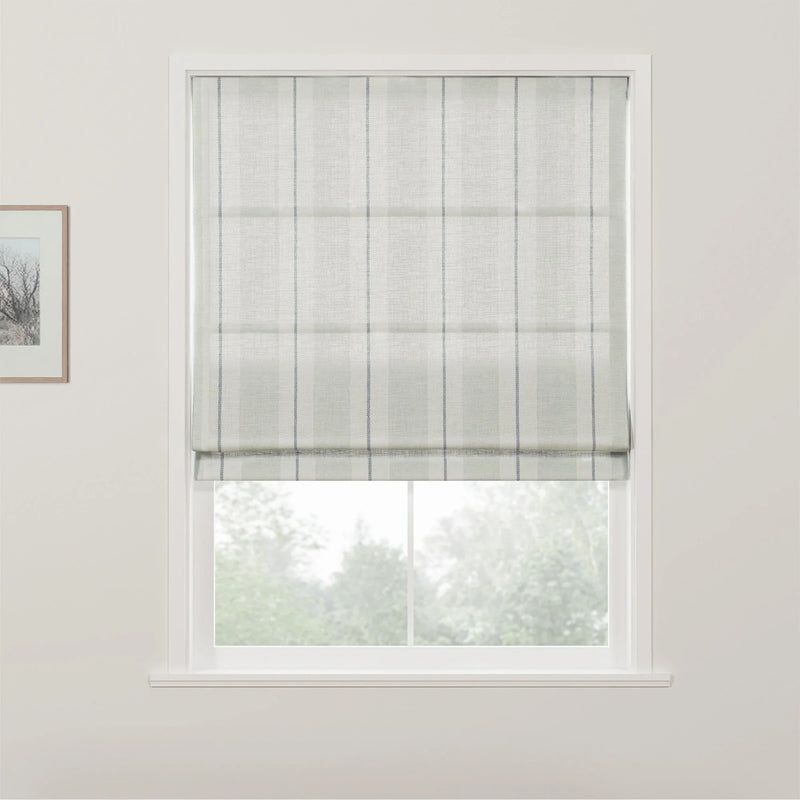 A light gray textured Roman blind with thin vertical blue stripes partially lowered in a white framed window offering a blurred view of green trees with a portion of a framed artwork visible on the left interior wall
