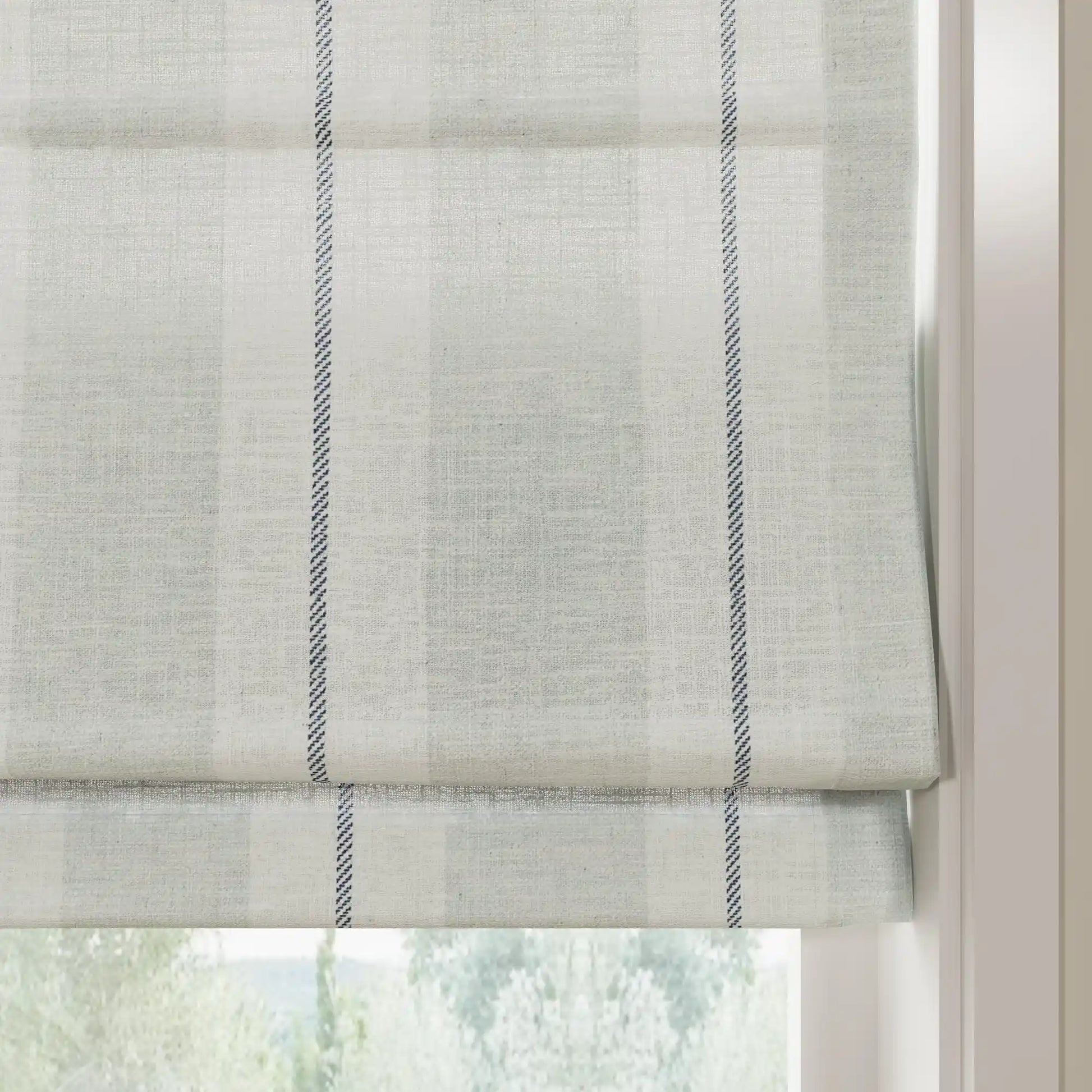 A light grey linen-blend Roman blind with a subtle plaid texture and prominent vertical dark blue dashed stripes, partially lowered in a window revealing a blurred green outdoor view